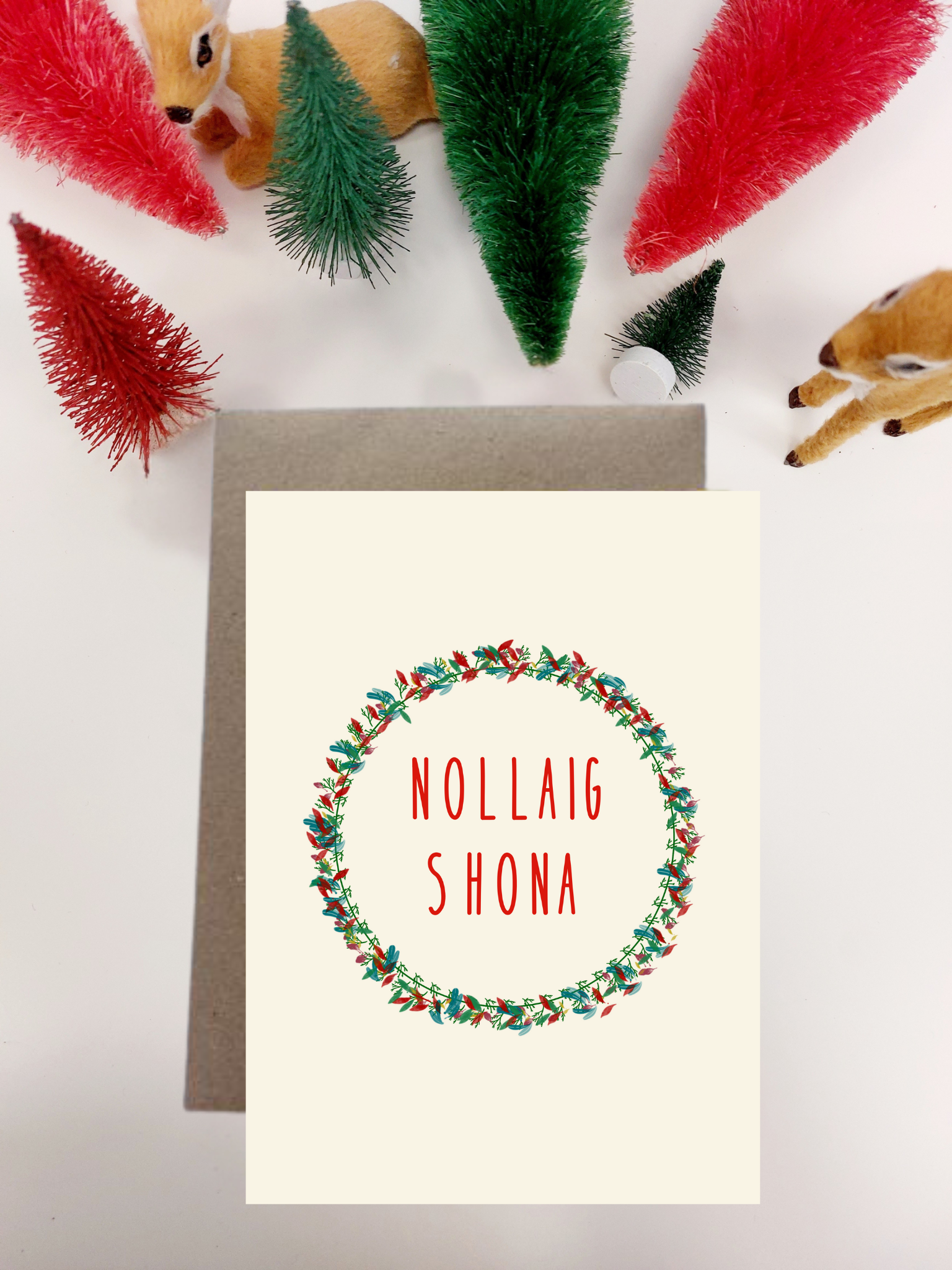 Irish Made Christmas card with wreath design and 'Nollaig Shona' text, surrounded by festive decorations on a white background.