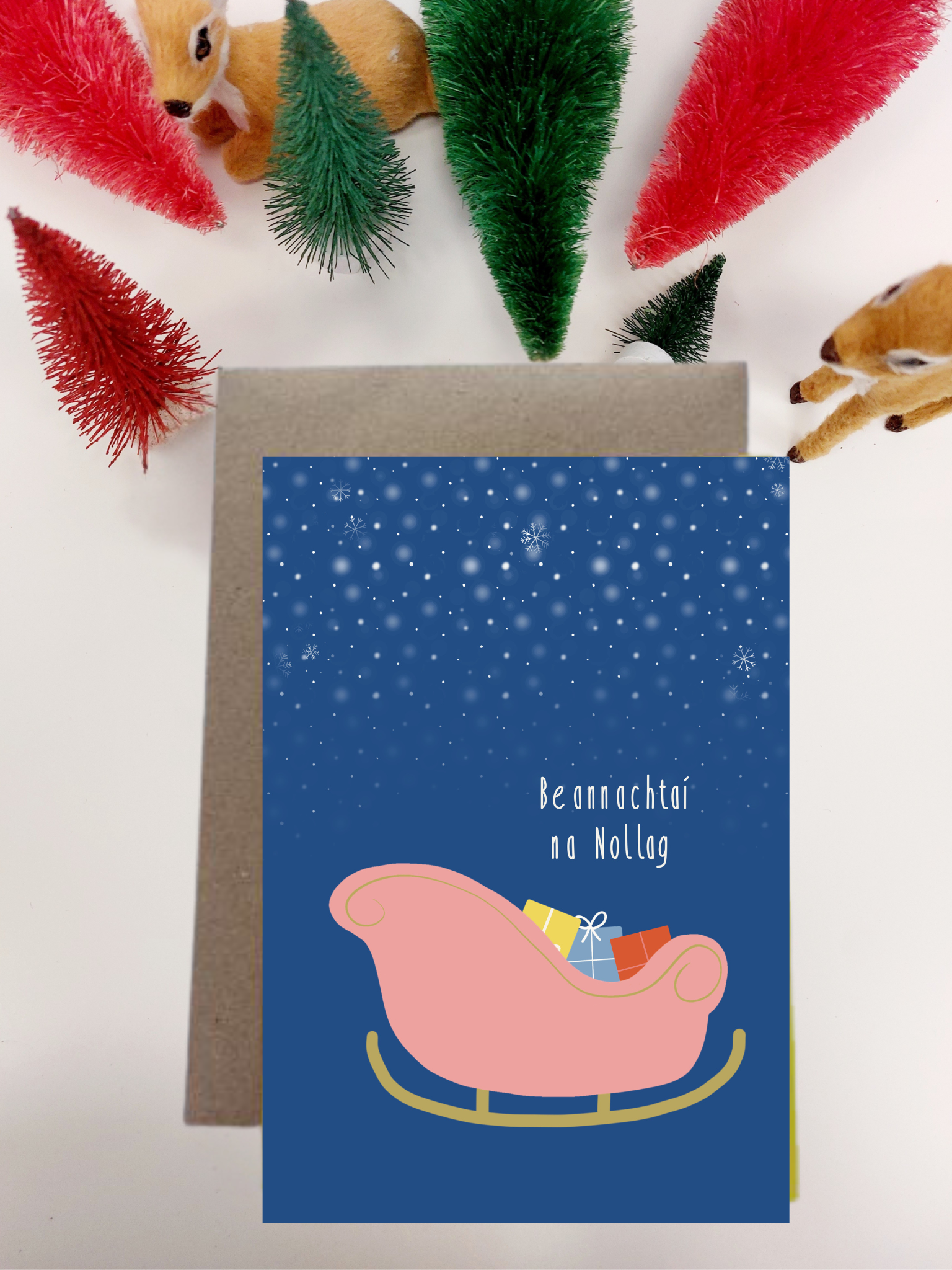 Irish lmade Irish language Greeting card with a pink sleigh and gift on a blue background, surrounded by festive decorations.