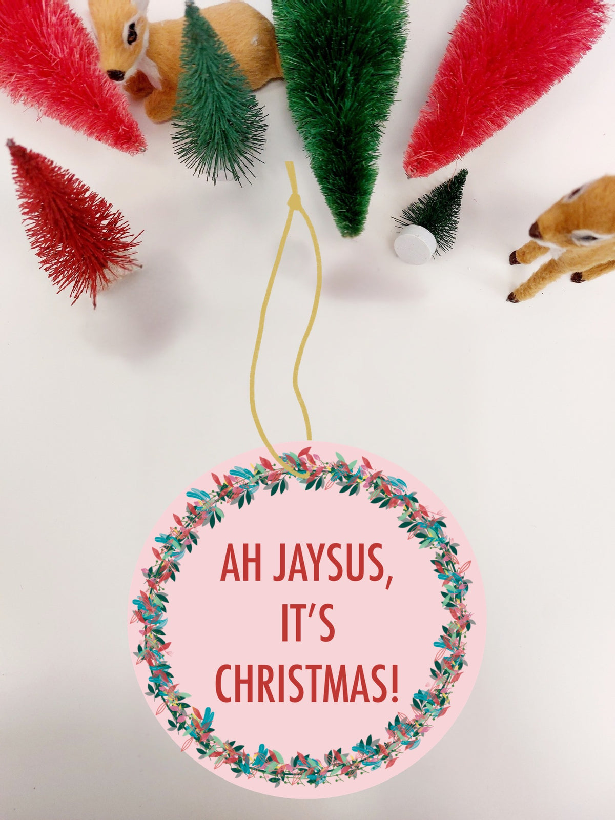 A round pink Christmas ornament with a floral wreath border and the text 'AH JAYSUS, IT'S CHRISTMAS!' in red, hanging from a yellow string. The background is white with festive props including small artificial Christmas trees and toy deer.
